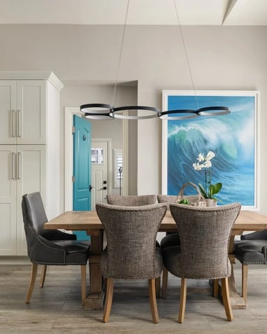 Open-plan kitchen and dining area with wood table, upholstered chairs, modern pendant light, and ocean wave artwork.