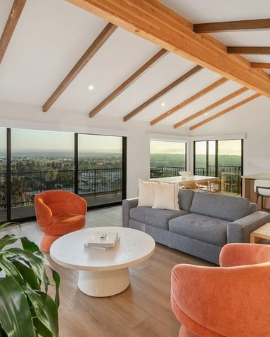 Modern open-plan living room with exposed wood beams, gray sofa, orange chairs, and floor-to-ceiling windows overlooking a city valley.