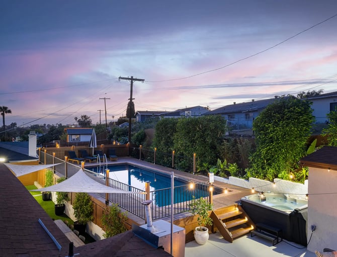 Backyard with illuminated pool, hot tub, and deck at dusk with mountain views and neighboring houses